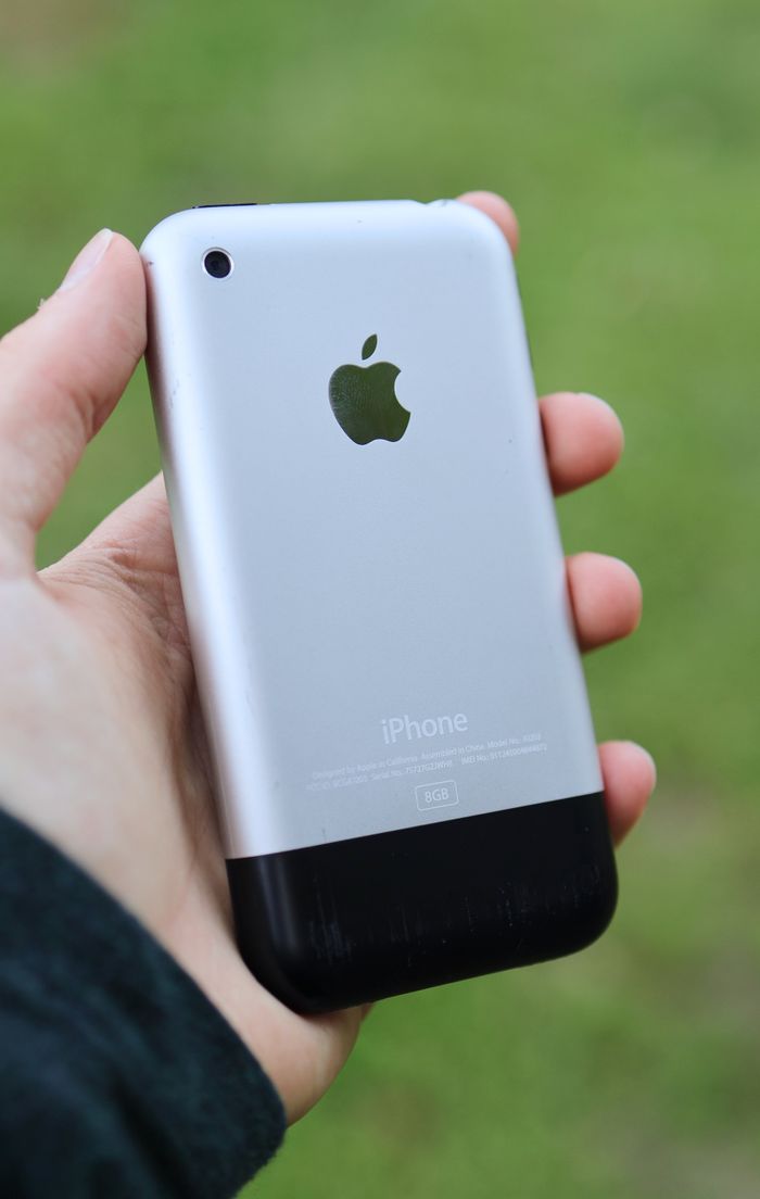 Close-up of a person holding a classic Apple iPhone outdoors on a blurred background.